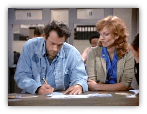 Man writing on paper with woman observing in a busy office setting.