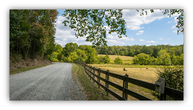 Gravel road alongside a wooden fence in a lush green landscape with trees and blue sky.