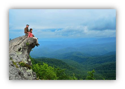 "Couple sitting on a rocky ledge overlooking a lush green mountain range under a cloudy sky."
