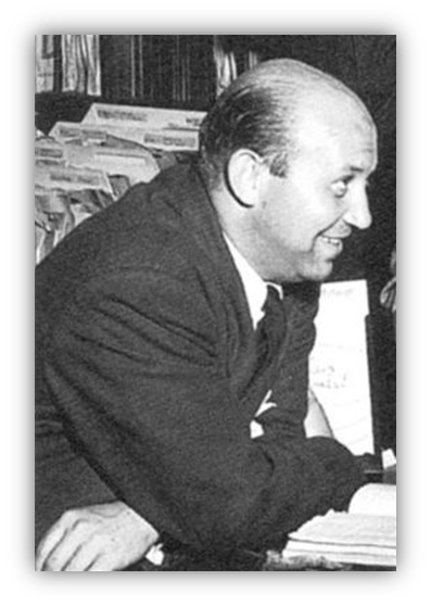 "Black and white photo of a smiling man in a suit, seated at a desk with papers."