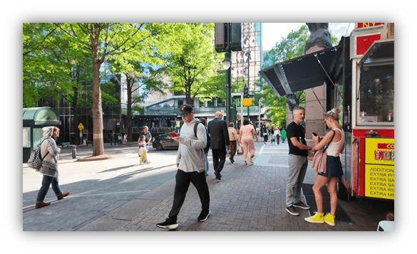 Busy city street with pedestrians and a food truck, surrounded by greenery.