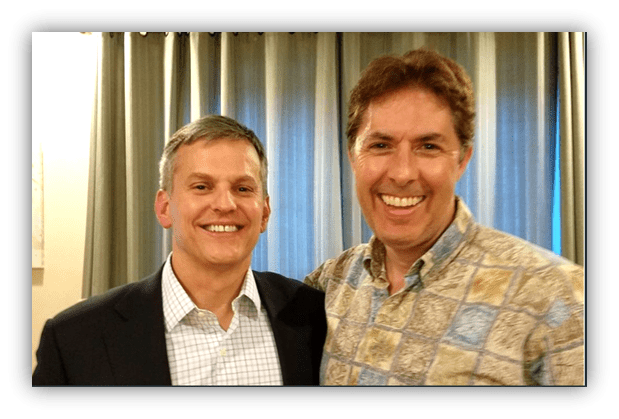 Two smiling men posing together indoors, with curtains in the background.