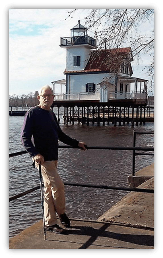 Elderly man with a cane standing near a lighthouse by the water.