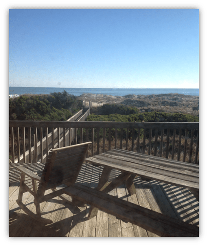 "Ocean view from a wooden deck with chairs overlooking sandy dunes."