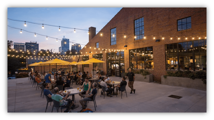 Outdoor dining scene with string lights and patrons enjoying food and drinks at a brick venue during sunset.