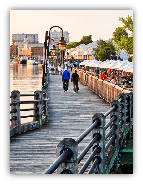 "Scenic waterfront boardwalk with people walking beside restaurants and sunset reflections."