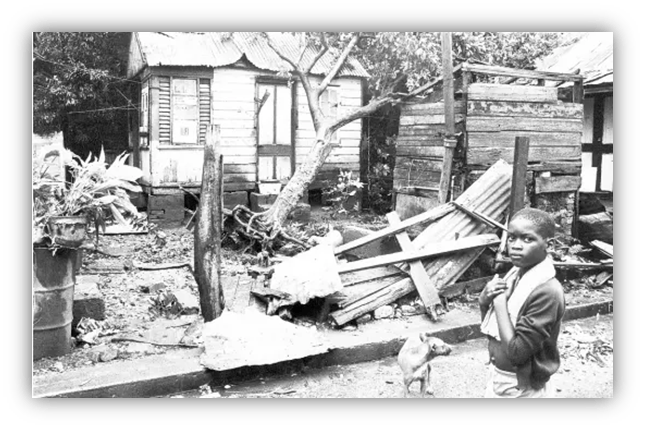"Black and white image of a boy standing near a damaged house and debris in a rural area."