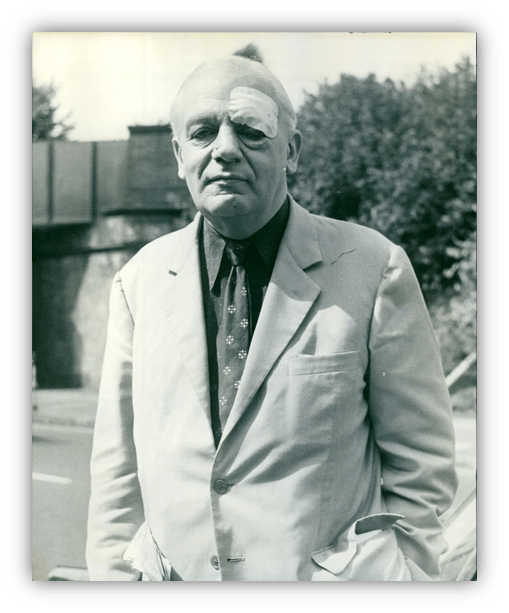 Black and white photo of a man with an eye patch, wearing a light suit, standing outdoors.