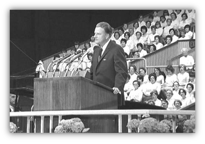 Billy Graham delivering a speech at a large event, with an audience in white attire.