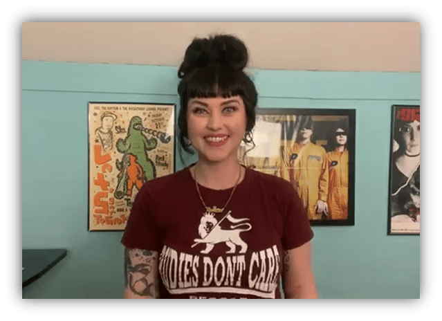 Smiling woman with tattoos wearing a "Hoodies Don't Care" t-shirt, standing against a colorful wall with vintage posters.