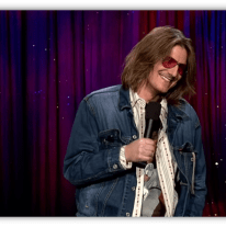 Stand-up comedian in denim jacket and sunglasses performing on stage.