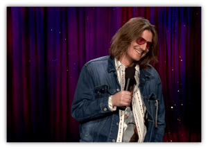 Stand-up comedian in denim jacket and sunglasses performing on stage.