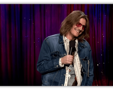 Stand-up comedian in denim jacket and sunglasses performing on stage.