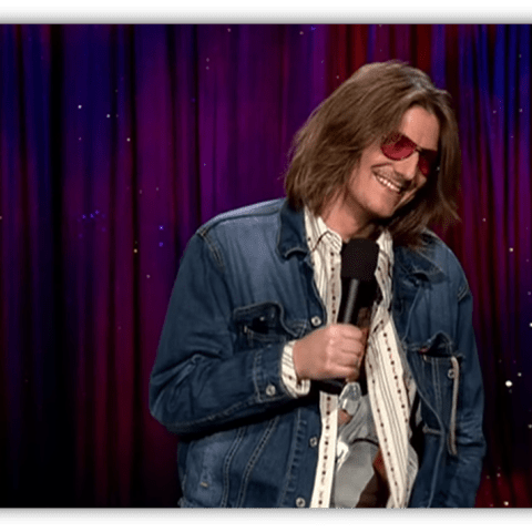 Stand-up comedian in denim jacket and sunglasses performing on stage.