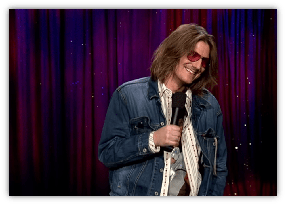 Stand-up comedian in denim jacket and sunglasses performing on stage.
