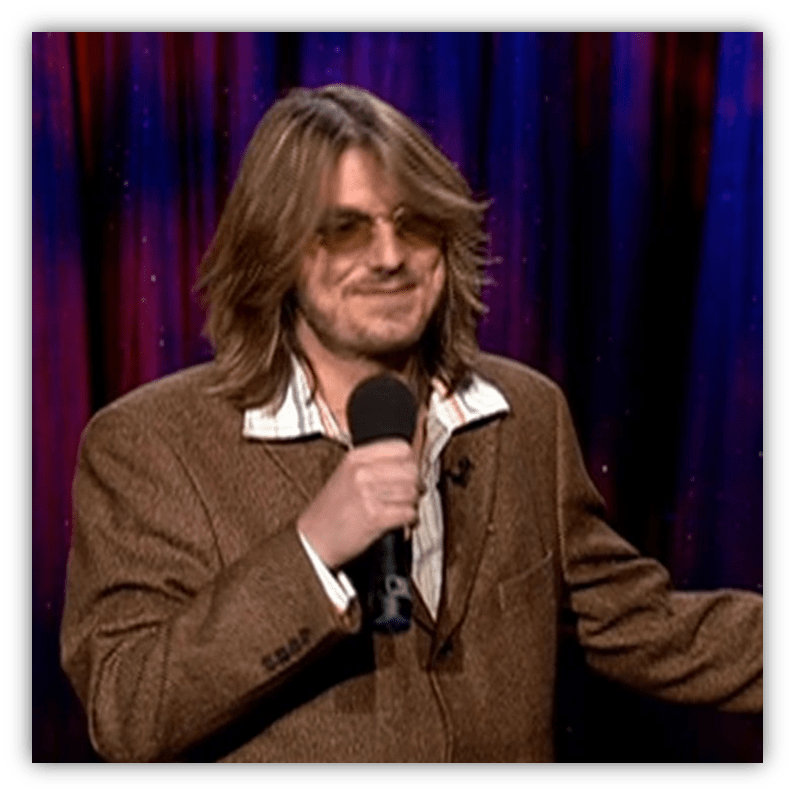 Comedian performing on stage with a microphone, wearing sunglasses and a brown jacket.