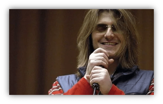"Smiling man with long hair holding a microphone on stage."