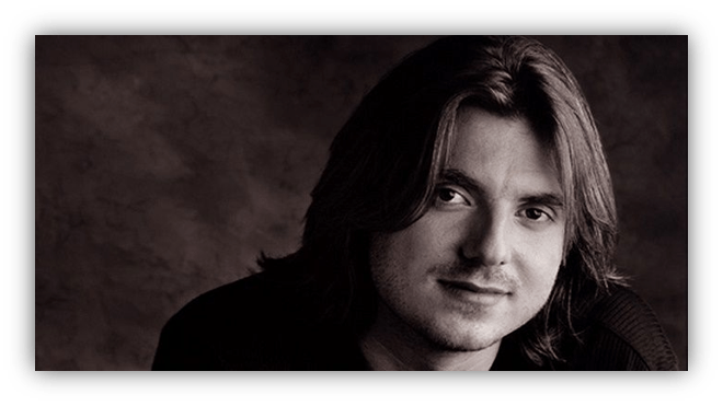 "Close-up portrait of a man with long hair, smiling against a dark background."