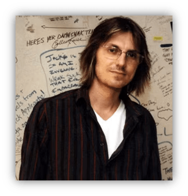 "Portrait of a man with glasses and long hair against a graffiti-covered wall."