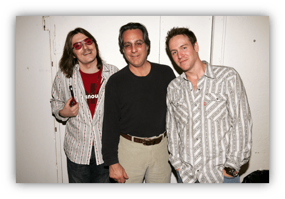 Three men posing together, dressed in striped shirts, smiling in front of a plain background.