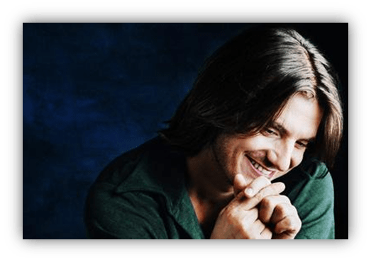 Smiling man with long hair in a dark blue background.