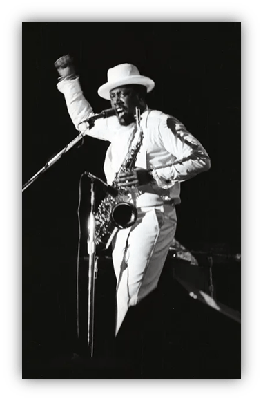 "Black and white photo of a saxophonist in a white suit and hat passionately performing on stage."