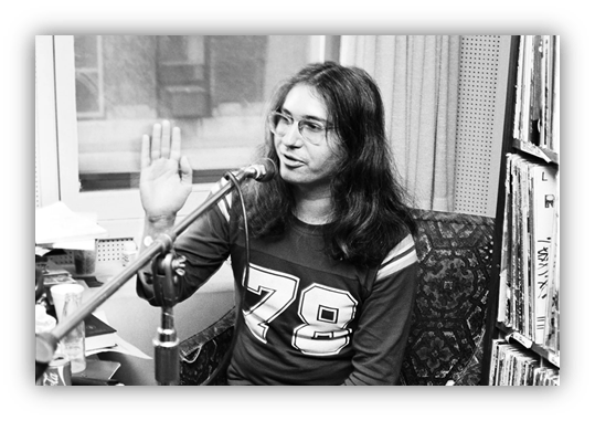 "Black and white image of a man with long hair and glasses speaking into a microphone, gesturing with his hand, surrounded by records."