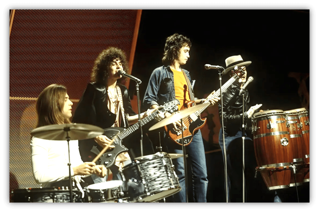 "1970s rock band performing live on stage with drums, guitars, and congas."