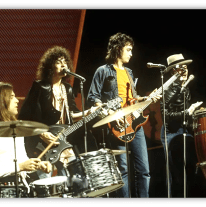 "1970s rock band performing live on stage with drums, guitars, and congas."
