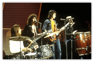 "1970s rock band performing live on stage with drums, guitars, and congas."