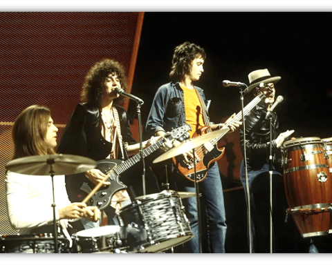 "1970s rock band performing live on stage with drums, guitars, and congas."