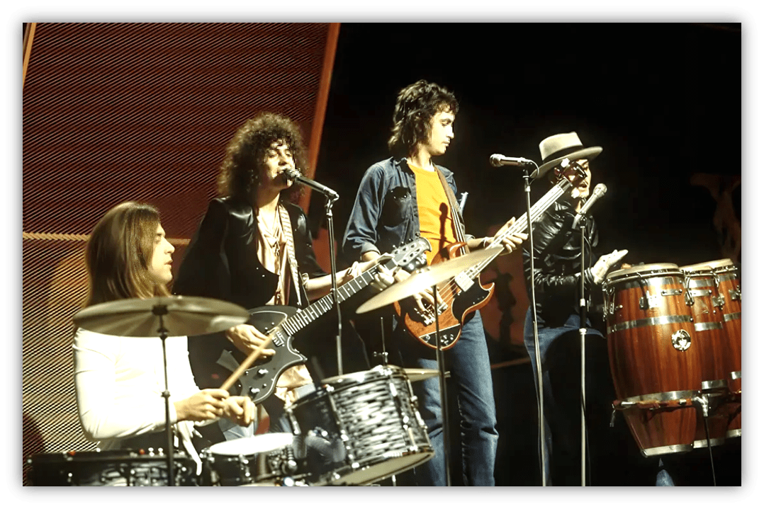 "1970s rock band performing live on stage with drums, guitars, and congas."