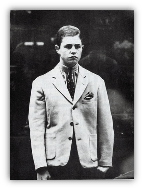 Black and white photo of a young man in a light suit with a polka dot tie and pocket square, standing solemnly.