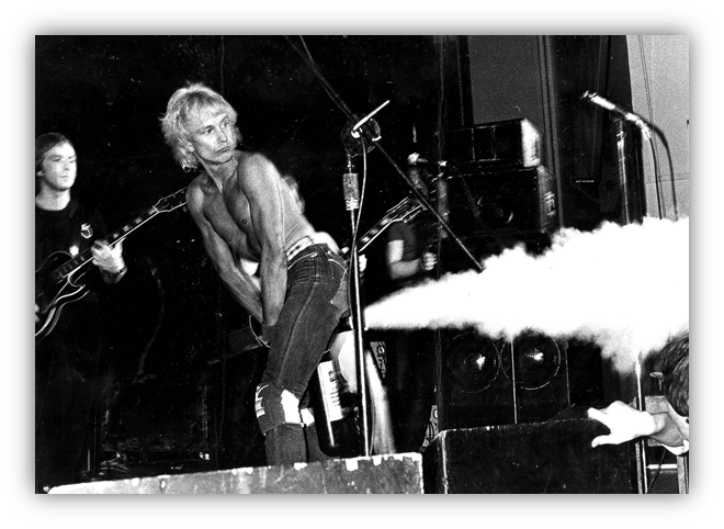 Rock concert performance with a shirtless vocalist and guitarist on stage, captured in black and white.