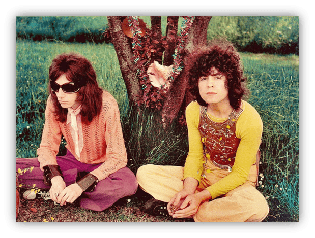 Two musicians in colorful 1970s outfits sitting by a tree in a grassy field.