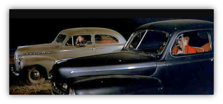 Classic cars parked at night with two people inside, one in each vehicle.
