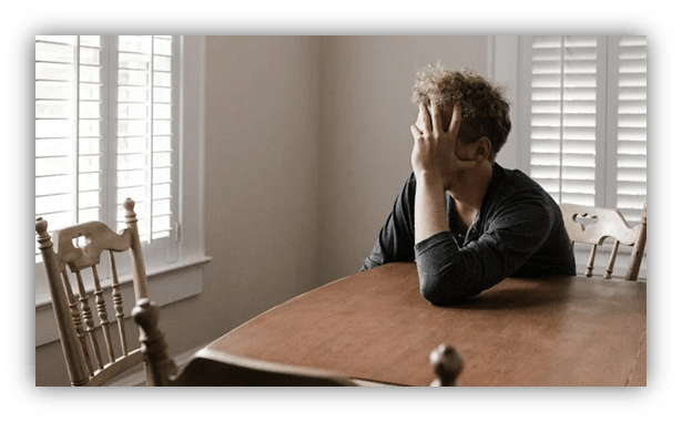 "Person with curly hair sitting at a table, looking pensive with hand on face."
