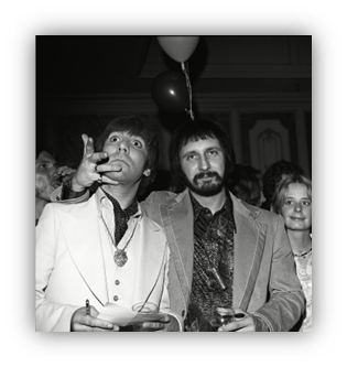 "Two men in vintage suits at a lively party, one holding a drink and gesturing."
