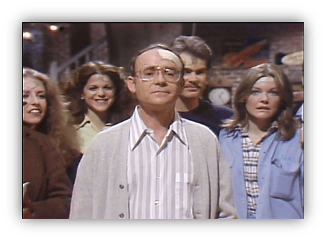 Group of six people, with a man in a cardigan and glasses in the foreground, smiling and looking at the camera.