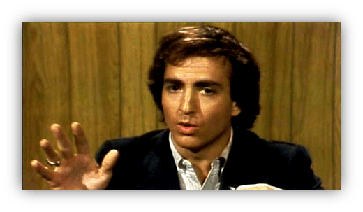 "Man speaking passionately at a desk with wooden backdrop."