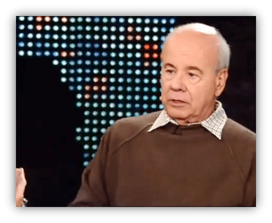 "Older man in a brown sweater speaking during an interview with a colorful backdrop."