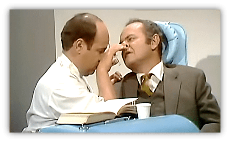 Two actors in a comedic scene, one examining the other's nose in a doctor's office setting.