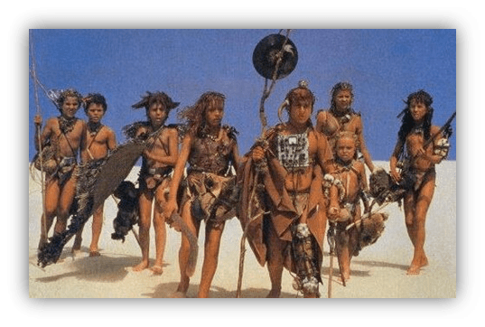 Group of children and adults in primitive attire holding weapons, set against a blue sky.