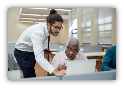 "Young man assisting elderly man with computer in a classroom setting."