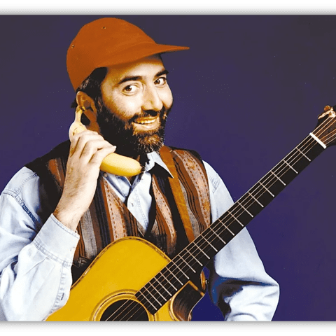 "Man in a cap holding a banana as a phone, smiling with an acoustic guitar."