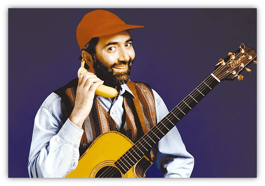 "Man in a cap holding a banana as a phone, smiling with an acoustic guitar."