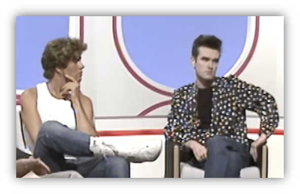 Two men sitting on a talk show set, one in a colorful jacket and the other in a tank top, engaged in conversation.