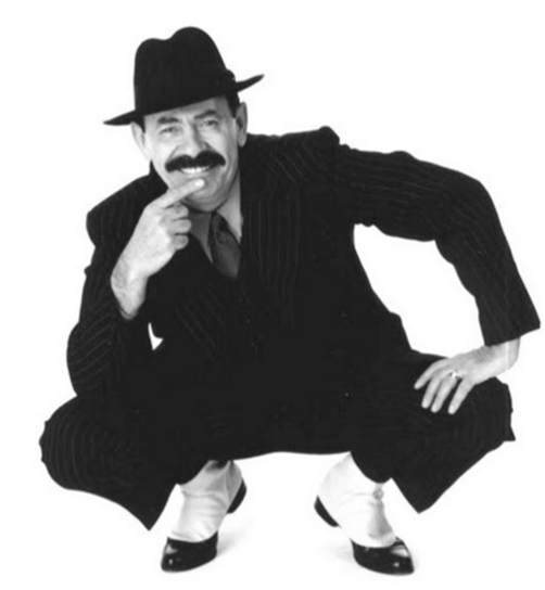 "Man in a stylish black suit and hat, striking a playful pose."