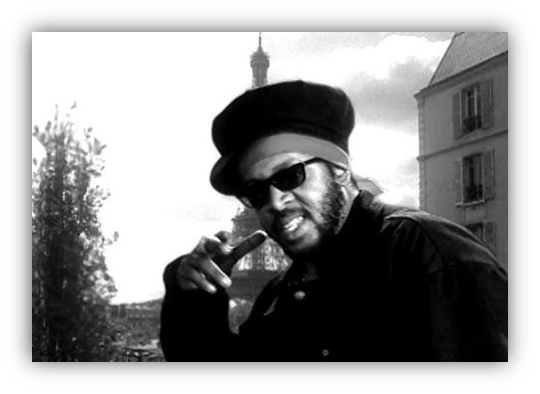 "Black and white image of a man in sunglasses and a hat, posing confidently with the Eiffel Tower in the background."