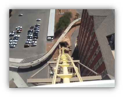 "View from the top of a roller coaster overlooking a parking lot and building."